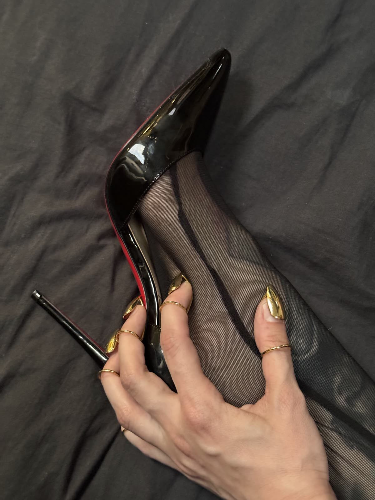 Aria's hand, gold rings and gold nail tips, holding a black patent stiletto.