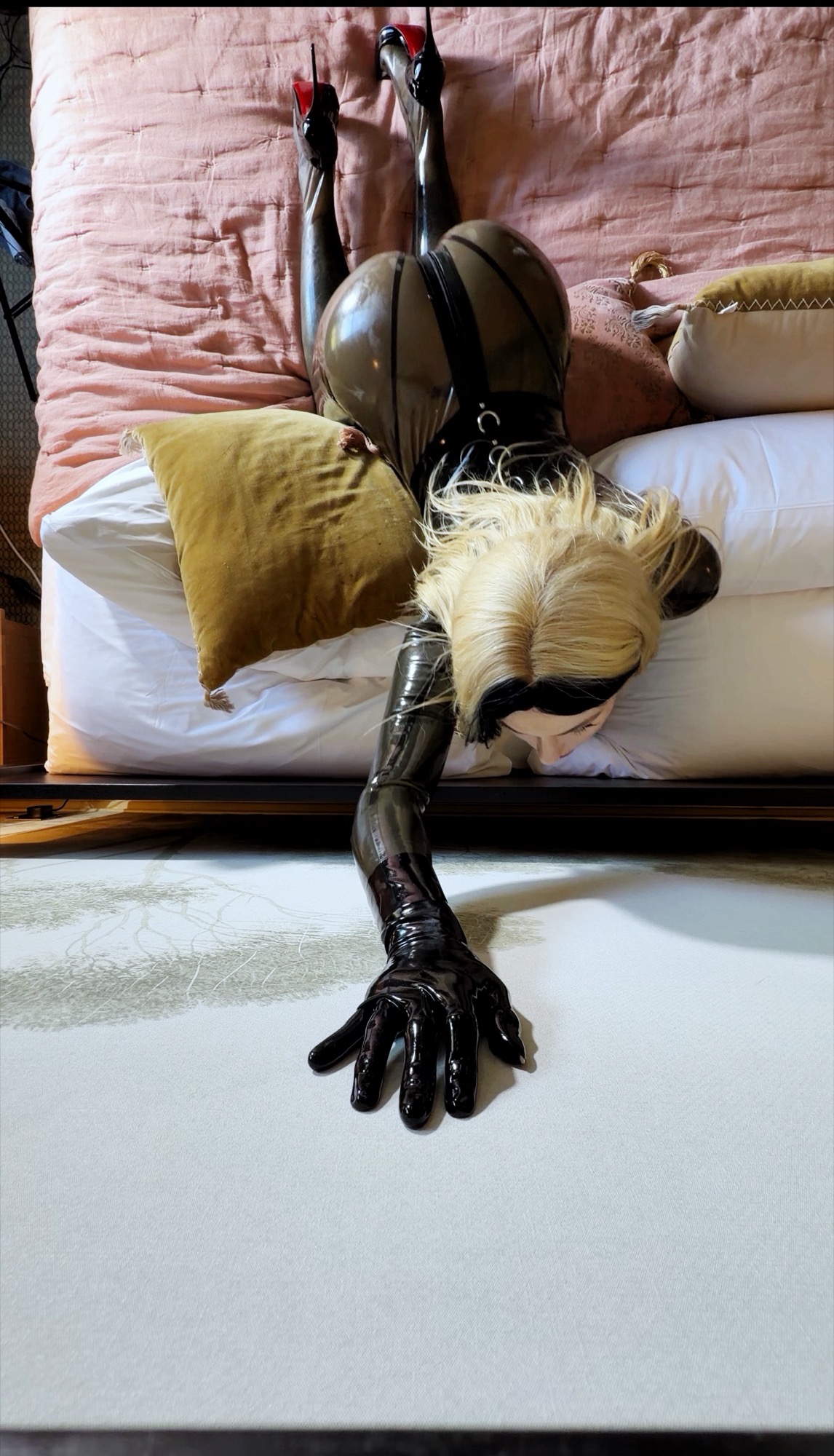 Aria in latex, reaching across a bed, photographed from above.