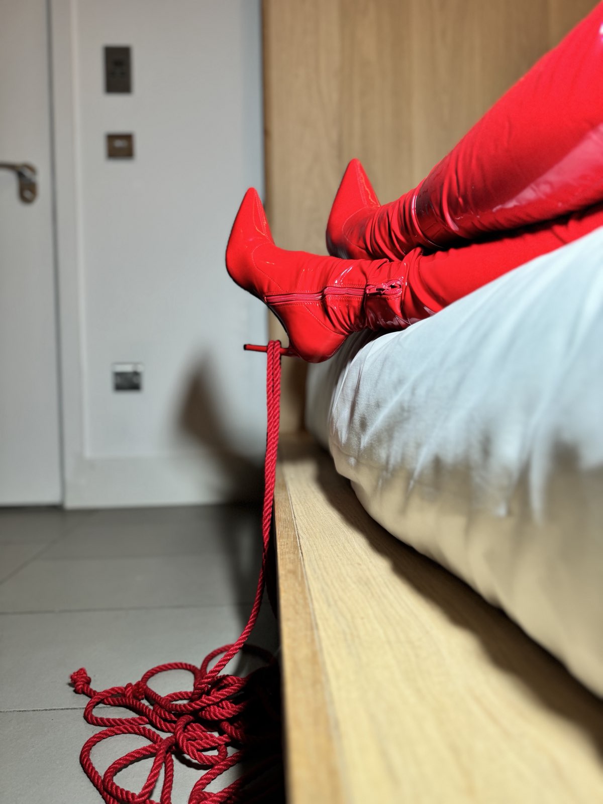 Aria in red patent thigh boots on a hotel bed, red bondage rope coiled on the floor beside her.