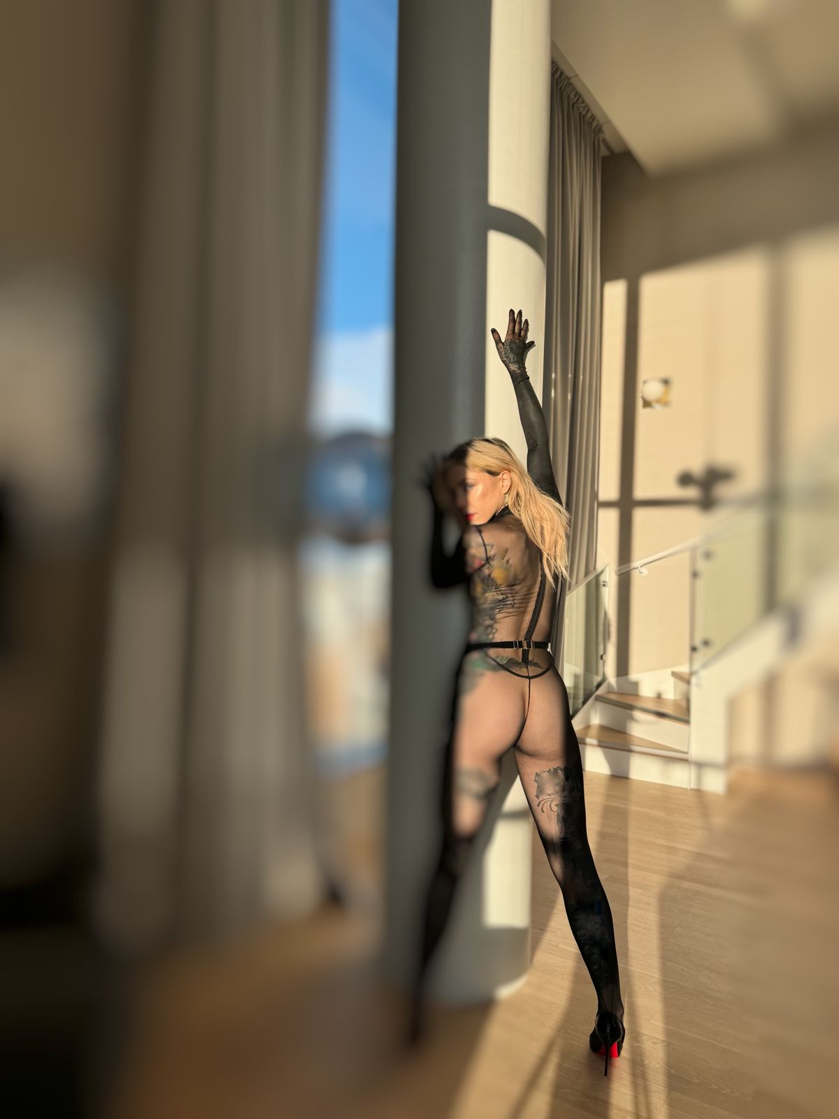 Aria in a sheer black bodysuit and heels, one hand raised against a sunlit window.